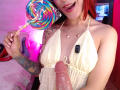 AmyStreetwise69 with red hair, holding a rainbow swirl candy, in a yellow halter top, showing a big, erect, circumcised penis, tattooed arm, and a necklace, smiling.
