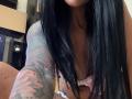AndraD has long black hair, visible tattoos on her right arm, and is wearing a pink bra, showing her small tits. Her skin is tan, and her mouth is slightly visible.