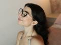 AnntonellaWatson with black glasses, wavy dark hair, pink lipstick, small gold earrings, white halter top, smiling, head tilted back, visible tattoo on right side of neck.