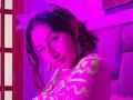 Photograph of MaddieLesYoux, face partially obscured by purple neon light, wearing a pink floral top, cigarette in mouth, suggestive gaze, dark hair, red window frame on left, sexual tension.