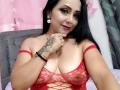 Zaria69 with black hair, red lace top, showing her tits, tattoos on hand, and smiling. She's wearing earrings, rings, and a bracelet. Pink and gray background.