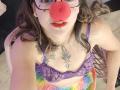 ArianaBlonde with a red clown nose, glasses, red lips, and a purple lace bra, showing colorful tie-dye pants and tattoos, looking up at the camera.