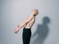 Topless EolantaMarina with blue eyeshadow, black pants, and tattoo on side, leaning back, arms crossed, shadow on wall, wearing gold bracelet, blonde hair in bun.