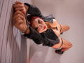 MaraCortes with red hair, black leather jacket, and fishnet stockings, arms cuffed, standing with hands above head, exposing her tits and bare ass, looking up with a smirk.