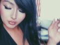 LilyConorr with glossy lips, closed eyes, and black hair, wearing a black top, has her hand up, looking down with a soft, sexy, slightly blurry, vintage-style photo.