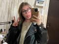 EleanoraMirabal with glasses, tongue out, black leather jacket, gold sweater, making a peace sign, phone in hand, light brown hair, black nails, in a bathroom mirror selfie.