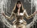 Tilliana stands center-frame, wearing a shiny, metallic gold dress with a deep V-neck and long sleeves. She has straight, medium-length brown hair with bangs, and wears round glasses. She holds a circular, crystal-encrusted frame with both hands, which is filled with water droplets. The metallic dress contrasts with the translucent, sparkling crystals. The image has a high-contrast, studio-lit look, emphasizing the texture of the dress and the clarity of the water droplets. The overall composition is balanced and symmetrical.