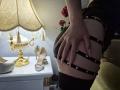 Rouzi's Hand umklammert ihren schwarzen, trägerlosen Hintern und enthüllt ihren nackten Po. Sie trägt schwarze Strumpfhosen und silberne High Heels stehen auf dem weißen Nachttisch. Im Hintergrund sind eine Lampe und Rosen zu sehen..
