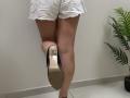 Tilliana's ass in white frayed denim shorts, standing on one leg with black heel, showing sole, green plant to right, white tiled floor, and light gray wall.