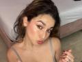 MilaGonz has pouty red lips, brown wavy hair, green eyes, and a gray tank top. She's on a tiled floor with a bed in the background.