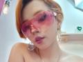 ChloeeWilliams with pink transparent goggles, orange bob, pink lipstick, gold earrings, and a gold necklace, showing her bare shoulders. Her pink-tinted goggles cover her eyes.