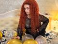 Niferttiti with long red curly hair, wearing a black fishnet top, sitting on a bed surrounded by gold and silver balloons, looking down with a subtle smile.