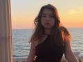 LeScharwath with wavy brown hair, wearing a black one-shoulder top, standing in a doorway at sunset, ocean in background, looking serious, soft evening light on her face.