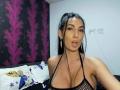 BrunetteBabe69 mit dunklem Haar, trägt ein schwarzes Netzoberteil, hat leicht geschwollene Lippen, trägt goldene Ohrringe und zeigt ihre kleinen bis mittelgroßen Brüste. Im Hintergrund ist eine Wand in Lila und Schwarz zu sehen..