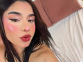 LunaVali met roze wangen, donkere wenkbrauwen, zwarte eyeliner-vleugels en rode lippenstift, met een frons en licht open mond, bruin golvend haar, een wit bed op de achtergrond..