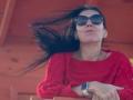 Nalini, con i lunghi capelli neri mossi dal vento, indossa occhiali da sole scuri, un maglione rosso e un orologio intelligente bianco, appare rilassata e sicura di sé..