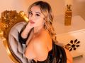 ZoeVibe with blonde hair, large tits, and a sexy smile, leaning on a gold chair, wearing a black leather top, exposing her breasts, in a warm, sexy, indoor setting.