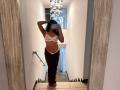 SofiaPassionne in a mirror selfie, wearing a white lace bralette, showing her small tits, and brown pants, with her hand in her curly black hair, in a white hallway.