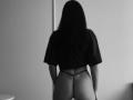 Black and white photo of Fridaa with long dark hair, facing away, wearing a black crop top and black thong, showing her ass, standing in a minimalist room.