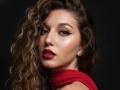 Sanella's face with wavy brown hair, red lipstick, and a red scarf, looking seductively at the camera with sultry green eyes and dark, well-defined eyebrows.
