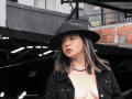 ArianaBlonde with black hat, black jacket, and chain necklace, showing small bare tits, has dark hair with blonde highlights, and a pouty expression, standing in an industrial setting.