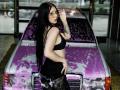 AlexLovely with long black hair, glasses, black bra, and lace pants, standing in front of a purple-splattered car, showing tattoo on right forearm, looking over shoulder.