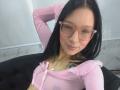 SophieDelaney with black hair, pink glasses, nose ring, pink sheer top, black cardigan, smiling, arm raised, visible tattoo on chest, pink lipstick, cute, sexy.