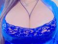 GilliSkquirtty's big tits in a blue lace bra, showing a lot of cleavage, with a silver necklace and a blue sheer robe, her long brown hair on the left.