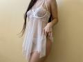 Tisia in a white lace and sheer babydoll, exposing her tits and pussy. Long dark hair, light skin, standing against a plain wall. Hand holding fabric.