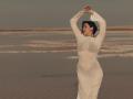 OliviaCluff in a sheer white dress, standing on a sandy beach, arms raised, eyes closed, with visible boobs and a curvy ass, dark hair, and a calm expression.