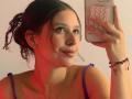 ValeryScot with dark curly hair in a bun, wearing a purple top, has cum on her chin, holding a pink phone with a circular button cover, smiling.