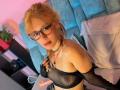 SuriEverly with blonde hair in a ponytail, wearing glasses, black mesh bra, and fingerless gloves, showing her tits, sitting on a bed with a dildo on the left.