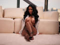 NyraNoble with long black hair, wearing an open white shirt, fishnet stockings, and no bra, sitting on a beige carpet, exposing her big tits and tattooed legs.