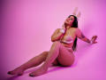 BonnieGray with big ass, white fishnet tights, bunny ears, and white bra, sitting on pink floor, hand on chin, tattoo on arm, looking up, pink lighting.