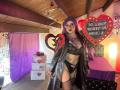 LilithDark69 with purple hair, glasses, black lingerie, garter, fishnet stockings, and black leather coat, standing in a room with heart decorations and a bed in the background.