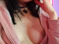IssisDiosa with black hair, wearing a pink zip-up hoodie, exposing her tits, small brown nipples, choker with a dark pendant, touching her lip, pinkish skin, dim lighting.