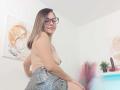 ArianaBlonde, topless with small tits, wearing checkered shorts, sitting, smiling, glasses, blonde hair, white walls, abstract paintings, potted plant, pink feathers, and a dildo in the background.