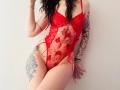 Tsukerberg in rode kant lingerie, met tatoeages op de armen en dij, staand tegen een witte muur, hand die donker haar aanraakt, rode strik aan de zijkant, slank, kleine borsten, zichtbaar schaamhaar..