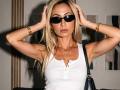 ViktoriiaLu with blonde hair, black sunglasses, white tank top, gold bracelets, and hands in hair, looking serious, holding black bag, gold necklaces, visible armpits.
