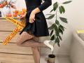 Rorria, vêtue d'une robe noire, la soulève pour montrer des collants résille et des talons hauts blancs, avec une jambe levée et une main sur ses fesses. Plante en pot et commode en bois..