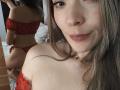 TaniaSandersX with red lace off-shoulder top and matching thong, showing her ass in mirror reflection, smiling at camera, long brown hair, fair skin, pink lips, necklace.
