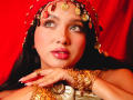 LilyConorr has shiny green eyes, glossy pink lips, and wears a gold coin headpiece with a red veil. Her hands are adorned with gold jewelry, resting on her chest.