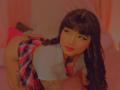 AnnieGrey with black hair, eyeliner, and red lipstick, wearing a plaid skirt and white top, on her knees, showing her tattooed arm and butt. Soft pink lighting.