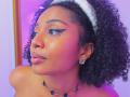 MeganWilliams has dark curly hair, a white headband, black eyeliner, glossy pink lips, a nose ring, and a necklace with black oval stones, wearing diamond earrings.