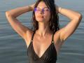 VikkiRi with wet black bikini top, arms up, hands behind head, purple mirrored sunglasses, wavy dark hair, tan skin, subtle makeup, calm water, bracelets, relaxed, confident expression.
