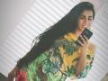AnnyFriend with long black hair, smiling, holding a black phone, wearing a yellow-green tropical-patterned shirt, one shoulder exposed, white blinds background, bright light, casual selfie, sexy, confident vibe.