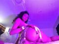 SophiaLuthor with curly hair, purple-lit, wearing a shiny, gold-embellished skirt, showing her ass, lying on a bed, looking at the camera, hand on her butt.