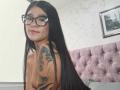 LuciadelaVega with long black hair, glasses, and tattoos on her shoulder, smiling at the camera, topless, showing small tits and a nose ring. Pink tufted headboard in background.