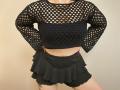 Niferttiti wears a black fishnet crop top with long sleeves and a black ruffled skirt, showing a tattoo on her right thigh. Boobs visible through the net.