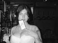 Black-and-white photo of Mettis with medium-length dark hair, holding a drink in one hand and a card in the other, wearing a white bra, showing tits and tattoos.